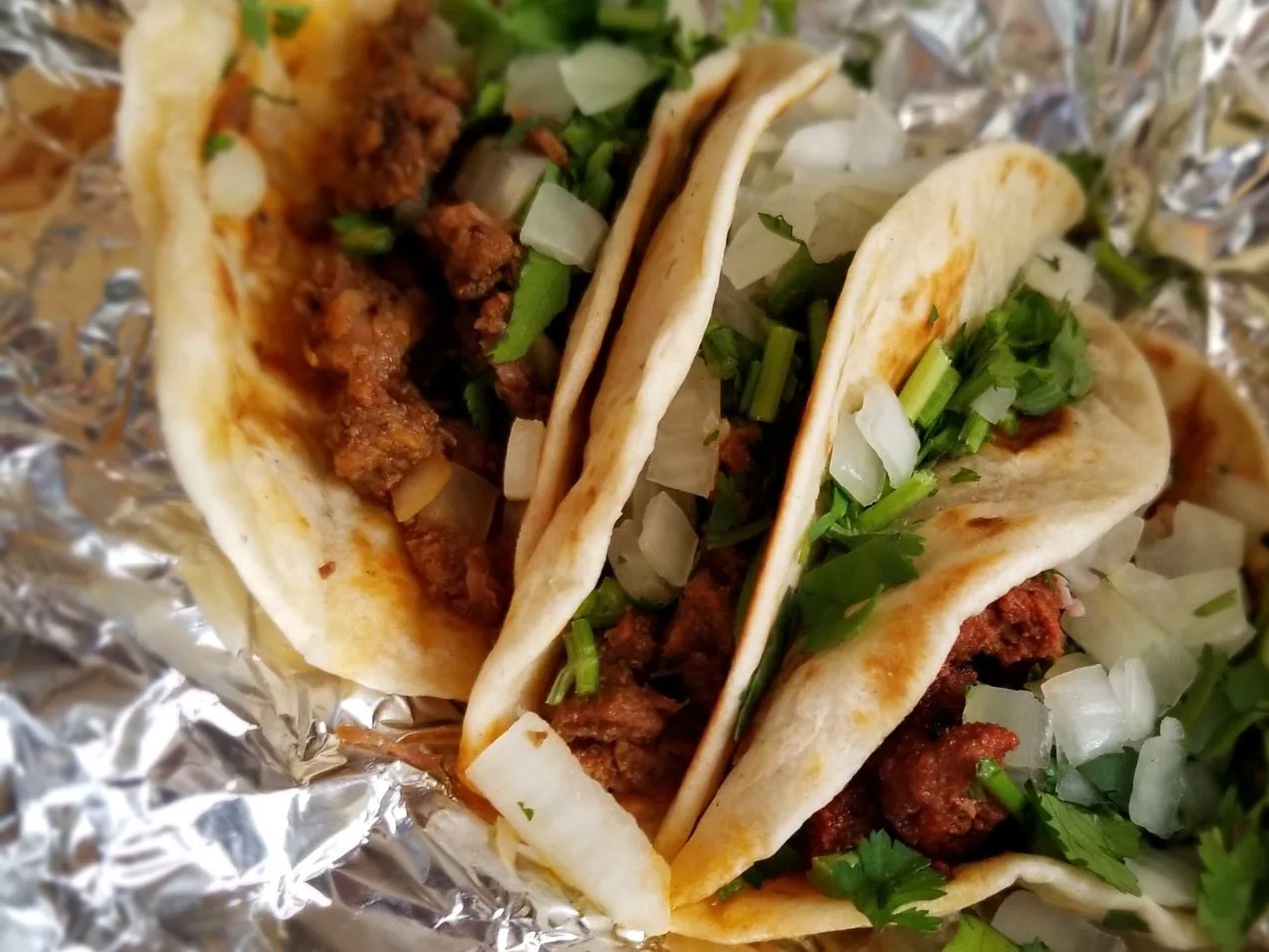 Three trompo tacos on foil at Tacos Tierra Caliente, topped with white onion and cilantro